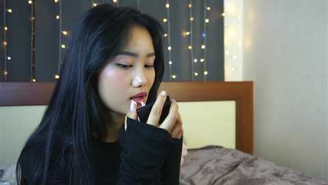 Media: Video of an East Asian woman with long black hair, wearing a black sweater, applying lipstick in a cozy bedroom with string lights and a gray bed.