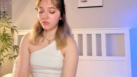 Media: Video of a young woman with light skin and shoulder-length brown hair, wearing a white one-shoulder dress, sitting on a white bed in a softly lit room.