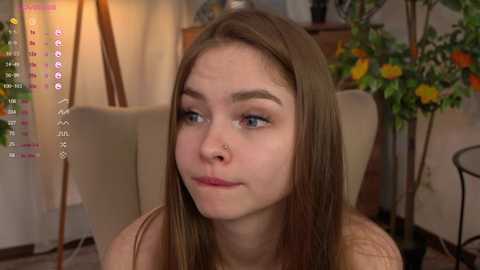 Media: Video of a young Caucasian woman with light skin, straight brown hair, and blue eyes, wearing a nose ring, sitting in a living room with a calendar and floral decorations.