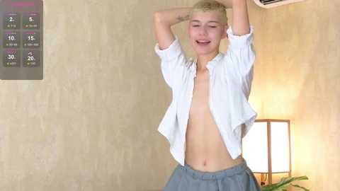 Media: Video of a smiling, fair-skinned, androgynous person with short blonde hair, wearing an unbuttoned white shirt and blue shorts, in a beige room with a thermostat and lamp.