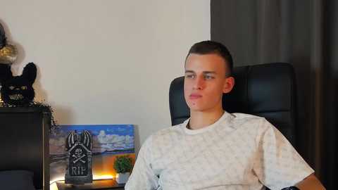 Media: Video of a young man with short brown hair, wearing a white hospital gown, seated in a black office chair, in a room with a black desk, a colorful painting, and a statue.