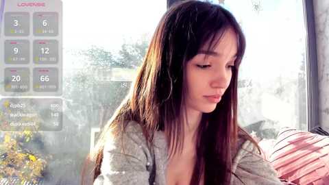 Media: A young woman with long brown hair, wearing a gray sweater, sits near a window, looking down thoughtfully. A virtual calendar and social media notifications overlay the scene.