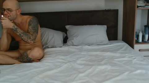 Media: Video of a bald, muscular tattooed man, nude, sitting on a white bed, holding a bottle, in a modern bedroom with dark headboard and nightstand.