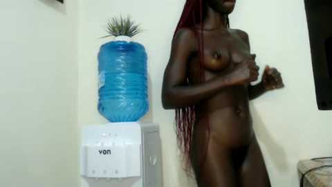 Media: Video of a nude, dark-skinned woman with long red braids, covering her breasts with her hands, standing next to a blue water dispenser and plant in a simple room.