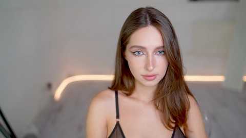 Media: Video of a young Caucasian woman with long brown hair and blue eyes, wearing a black lace bra, standing in a modern, dimly lit room with a sleek, white sofa.