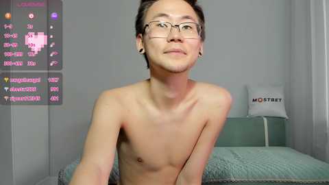 Media: Video of a shirtless, slender Asian man with glasses, sitting on a bed in a minimalist room with a green quilt, a white pillow, and a \"MOBET\" pillow.