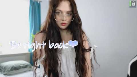 Media: Video of a young woman with long brown hair, wearing glasses and a white top, standing in a bright, modern room with a bed and blue curtains in the background. Text overlay reads, \"Be right back,\" with a blue heart emoji.