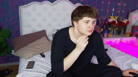 Media: Video of a young boy with straight brown hair, sitting on a bed with white bedding, purple wallpaper, and a floral arrangement in the background.