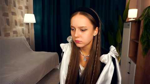 Media: Video of a young girl in a French maid costume, with long brown hair, in a dimly-lit room with a white bed, dark curtains, and potted plants.