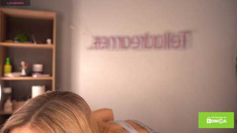 Media: A blurry photo of a woman's back in a white bra, with \"#amorebulllet\" graffiti on a beige wall. Shelves with green bottle, red cup, and white container in the background.