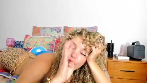 Media: Video of a blonde woman with curly hair, lying on a colorful bed, wearing a blue bikini, touching her face. Background includes a wooden dresser with a camera and a cup.