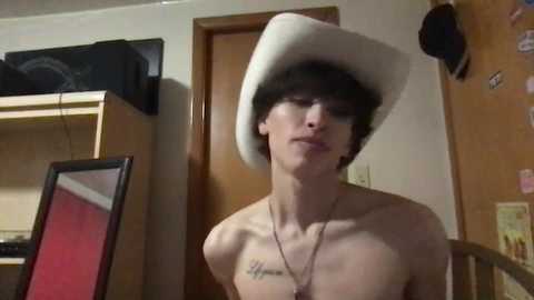 Media: Video of a slender, shirtless young man with short dark hair, wearing a white cowboy hat, standing in a cluttered room with wooden furniture, a mirror, and a TV.