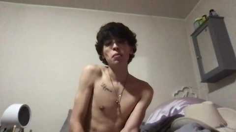 Media: Video of a shirtless, young, slender male with short, curly hair and a small tattoo on his chest, sitting on a bed with messy sheets in a dimly lit room.