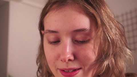 Media: Close-up video of a young Caucasian woman with wavy, light brown hair and fair skin, eyes closed, looking serene, with a soft, neutral background.