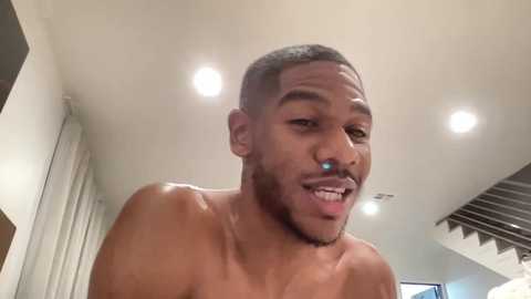 Media: Video of a shirtless, muscular Black man with short hair, light skin, and a trimmed beard, smiling, in a modern, well-lit room with white walls and recessed lighting.