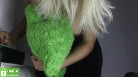 Media: Video of a blonde woman with long hair in a black tank top holding a bright green, textured pillow. She's in a dimly lit room with a gray wall.