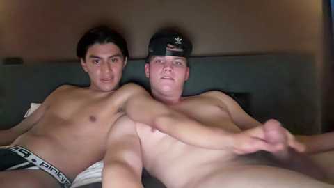 Media: Video of two shirtless, young, muscular men lying on a bed, wearing Calvin Klein underwear. One has a black cap, the other has a tattoo. Background is dimly lit, with a neutral-colored wall.