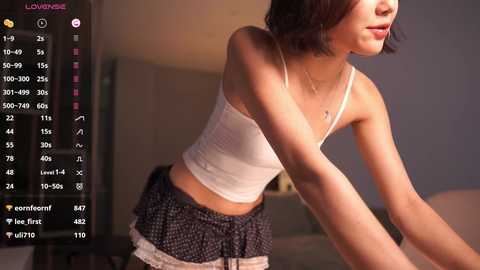 Media: Video of an Asian woman with short dark hair, wearing a white tank top and black polka-dot skirt, leaning forward, in a dimly lit room with a digital clock and heart icons.