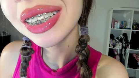 Media: Video of a light-skinned woman with glossy red lipstick, braid hairstyles, and metal braces, wearing a pink sleeveless top. Background shows a white shelf with various items.