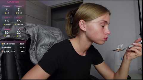 Media: Video of a blonde woman with light skin, wearing a black t-shirt, holding a makeup brush. She's indoors, seated, with a grey jacket draped over a chair. Live streaming video overlay on the left.