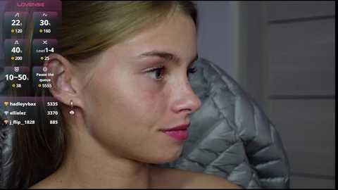 Media: A video shows a young, light-skinned woman with long, straight blonde hair and blue eyes, wearing a silver earring, sitting against a gray quilted pillow. She has a neutral expression.