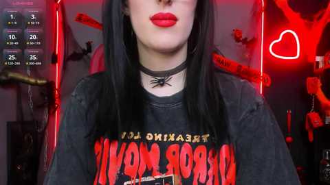 Media: Video of a woman with pale skin, long black hair, wearing a black choker, red lipstick, and a \"Marilyn Monroe\" graphic tee in a dimly lit room with red neon lights, black spider web accents, and BDSM paraphernalia.