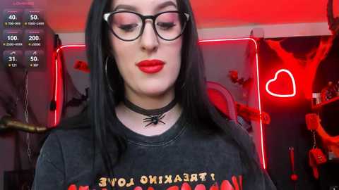 Media: Video of a pale-skinned woman with long black hair, wearing black-framed glasses, a black choker with a spider design, and a gray T-shirt with \"HALLOWEEN\" in red letters. Background features red neon lights, bat decor, and a heart-shaped light.