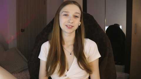 Media: Video of a young, fair-skinned woman with long brown hair, wearing a white, button-up top and silver earrings, seated in a plush, black chair in a softly lit bedroom.