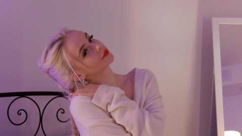 Media: Video of a slender, blonde Caucasian woman with pale skin, wearing a white off-shoulder sweater, leaning against a black wrought iron bed frame in a softly lit, minimalist room.