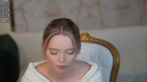 Media: Video of a fair-skinned, blonde woman with loose hair, wearing a white off-shoulder top, sitting on a vintage gold-framed chair with a green cushion, in a softly lit, beige-walled room.