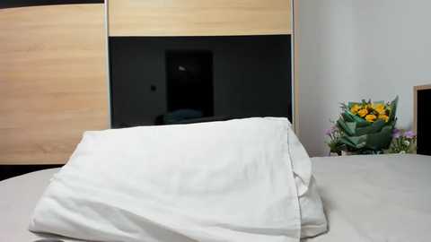 Media: Video of a bed with a white pillow on a light grey duvet, wooden wardrobe with a black door in the background, and a bouquet of yellow and purple flowers on the right.