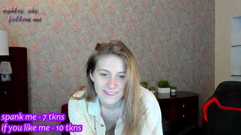 Media: Video of a young woman with light brown hair and a casual outfit, sitting in a room with floral wallpaper and a gaming chair in the background.