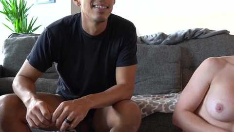 Media: Video of a smiling, shirtless, muscular man of mixed ethnicity with short hair, wearing shorts, sitting on a couch with a partially nude woman, topless with small breasts, both facing each other.