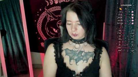 Media: A young woman with pale skin, long black hair, and a black choker with a pentagram, wearing a black lace top, sits in a dimly-lit room with dark walls and a glowing pentagram.