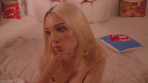 Media: Video of a blonde woman with long hair, fair skin, and large breasts, biting a lollipop, wearing a red bra. She is indoors on a bed with white sheets, surrounded by colorful pillows and a blue and red box.