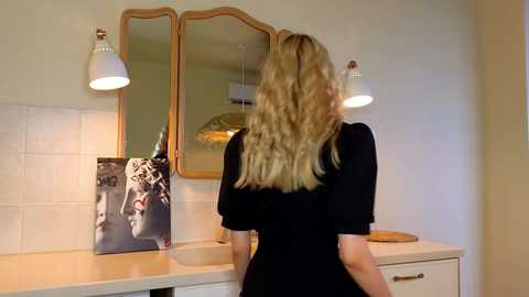 Media: Video of a blonde woman with wavy hair, wearing a black dress, standing in a modern bathroom with white tiles, a white countertop, and a framed photo of a woman on the left.