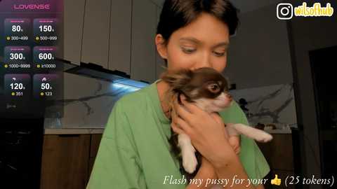 Media: Video of a young woman with short black hair, wearing a green shirt, holding a small brown and white Chihuahua pup, in a modern kitchen.