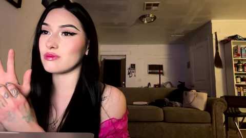 Media: A video of a young woman with long black hair, wearing a pink off-shoulder top, making a pouty face in a cozy, dimly lit living room with a beige couch and bookshelf.