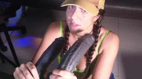 Media: Video of a young woman with light skin and long brown hair in braids, wearing a yellow cap and green tank top, kneeling in a dimly-lit room, sucking on a large, black dildo.