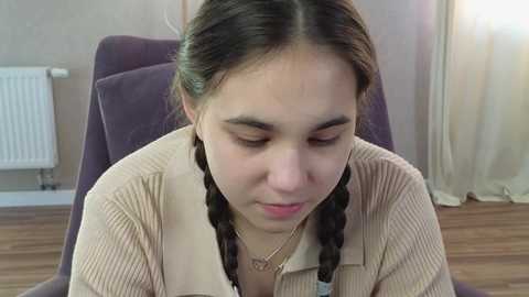 Media: Video of a young girl with long brown hair in braids, wearing a beige sweater, sitting on a dark chair in a room with a radiator and curtains.