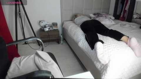 Media: Video of a sleeping woman in black pajamas on a white bed with a quilted headboard, surrounded by a minimalistic bedroom with white walls, a lamp, and a chair.