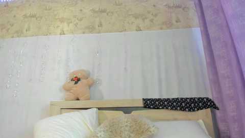 Media: A video of a simple bedroom featuring a beige teddy bear on a white bed with a patterned pillow, a black and white polka-dot scarf draped over the headboard, and light purple curtains.