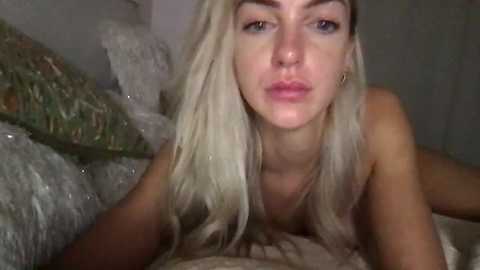 Media: Video of a topless, fair-skinned woman with platinum blonde hair, lying on a bed with patterned bedding, making a pouty face.