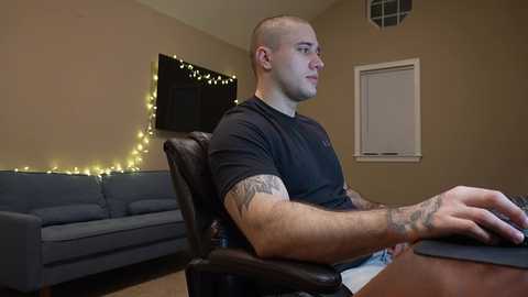 Media: Video of a muscular, bald man with a tattooed arm, wearing a black t-shirt, seated in a black leather chair, typing on a computer in a beige room with a gray couch, string lights, and a dark TV.