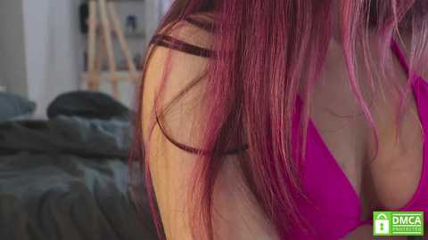 Media: Video of a woman with long, vibrant red hair, wearing a pink top, lying on a bed with messy gray sheets in a dimly lit room.