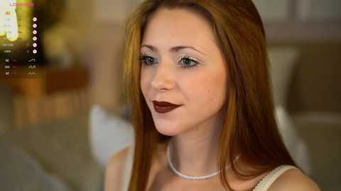 Media: Video of a fair-skinned woman with long, straight auburn hair, wearing a white top and a pearl necklace, smiling gently. Background is softly blurred, featuring a wooden table and a cozy, warm-toned room.