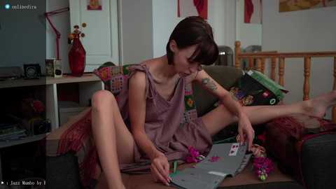 Media: Video of a slender Asian woman with short brown hair, wearing a mauve dress, seated on a couch, playing cards, in a cozy living room with wooden furniture and colorful decor.