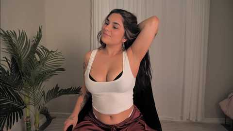 Media: Video of a Latina woman with long, dark hair, wearing a white tank top and red pants, posing with her hand behind her head in a cozy indoor setting.