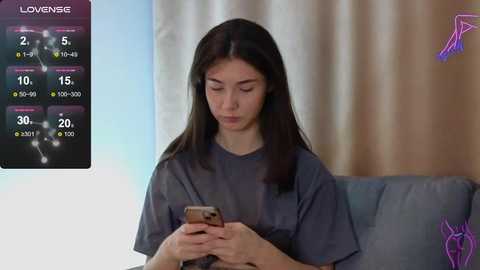 Media: Video of a young Asian woman with long dark hair, wearing a gray shirt, looking at her smartphone in a living room with beige curtains and a gray couch.