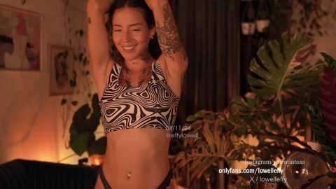 Media: Video of a smiling, tattooed woman in a black and white zebra-print sports bra, stretching in a dimly lit room with green plants and framed art.